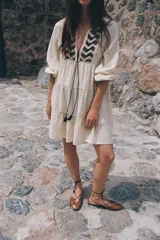 Vestido corto color crudo con escote en pico con cordón con borlas y manga larga. Presenta bordados en el frente.