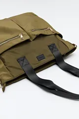 Bolso tote marrón de nylon con doble asa de mano negra y correa bandolera negra regulable y desmontable. Tiene dos bolsillos frontales con cierre metálico dorado.