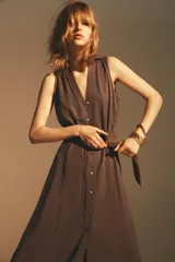 Vestido midi marrón sin mangas, con cuello solapa, escote en pico, cierre frontal con botones y cinturón de la misma tela con hebilla metálica.