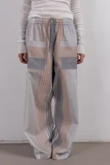 Pantalón ancho con cintura elástica y cordón ajustable, bolsillos frontales y diseño patchwork con rayas verticales y horizontales en tonos neutros.