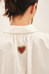 Camisa blanca de algodón con bordados de corazones rojos y dorados en el delantero. Tiene cartera en el delantero y fruncido en el centro de la espalda.