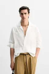 Camisa blanca de corte relaxed fit confeccionada en tejido de algodón y lino. Tiene cuello bowling, manga larga con puños abotonados, jaretas en el delantero, bolsillos de parche en pecho y cadera, bajo con aberturas laterales y cierre frontal con botones.