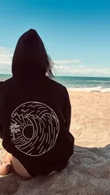 Hoodie negro con capucha y cordones ajustables. Estampado en la espalda con un diseño circular que incluye una ola, un sol y una palmera.