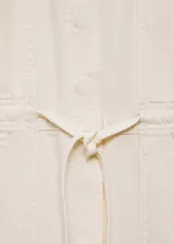 Mono largo color crudo, sin mangas, con cuello camisero, cierre frontal con botones, cintura ajustable con lazo y bolsillos delanteros y traseros. Diseño recto.