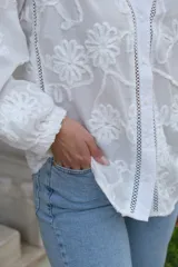 Camisa blanca con bordado de flores, cuello en V, cierre frontal con botones, mangas largas con puños elásticos y detalles de encaje.