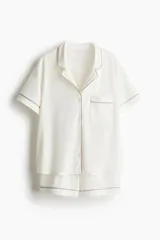 Conjunto de pijama corto color crema, compuesto por una camisa de manga corta con cuello abierto, botones al frente, bolsillo superior y ribetes en color marrón, y un short con cintura elástica y ribetes en color marrón.