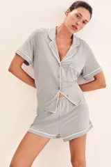 Pijama corto de dos piezas color gris con ribetes blancos. Camisa de manga corta con cuello abierto, botones adelante, hombros caídos y bolsillo superior. Short con elástico en la cintura.