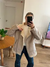 Campera gamulán color beige con cuello, solapas y puños de piel sintética. Cierre frontal con cremallera asimétrica y hebilla en el cuello.