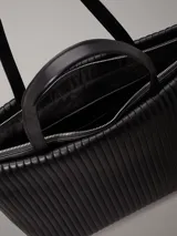 Bolso tote negro Calvin Klein, confeccionado en piel sintética acolchada con diseño de rayas verticales. Tiene doble asa de hombro, cierre superior con cremallera, herrajes plateados y logo metálico en el frente.