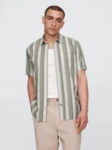 Camisa de poplin a rayas verticales en tonos verde oliva y blanco, de manga corta y corte recto. Tiene cuello clásico, cierre con botones y bolsillo en el pecho.