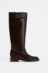 Bota alta de cuero color marrón oscuro, con caña alta, punta redonda y hebilla decorativa en la parte superior.
