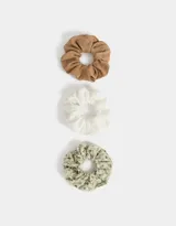 Set de tres scrunchies para el pelo con diferentes estampados: uno liso color marrón, uno blanco con bordado calado y uno con estampado floral en tonos verdes.