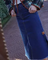 Falda larga de jean azul oscuro, corte amplio y recto, con abertura frontal y terminación desflecada en el ruedo. Presenta un pequeño parche de cuero marrón en la abertura lateral.