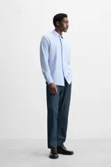 Camisa celeste a rayas verticales blancas, de corte recto, con cuello italiano abierto, manga larga con puños abotonados y cierre frontal con botones.