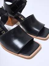 Sandalias de cuero negras con punta cuadrada, tira ancha sobre el empeine y pulsera con hebilla dorada al tobillo. Taco geométrico de madera clara.
