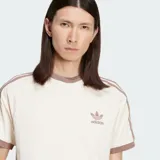 Remera Adidas color beige con cuello redondo y mangas cortas con ribete marrón. Presenta las tres rayas características de la marca en color marrón en los hombros y el logo bordado en el pecho.