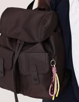Mochila color crema de tela acolchada con pespuntes verticales. Tiene un bolsillo frontal con cierre, doble cierre superior, manija superior, correas regulables y bolsillos laterales. Incluye un charm colgante de corazón.