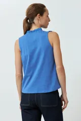 Top blanco sin mangas, con cuello alto y volados en los hombros. Presenta detalles de broderie en el cuello y el pecho, y cierre de botones en la parte superior delantera.