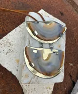 Collar con colgante de dos piedras naturales irregulares engarzadas en metal plateado y dorado, suspendidas de un cordón de cuero marrón con detalle de nudo corredizo. Las piedras tienen vetas en tonos naranja, marrón y blanco.