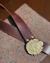 Cinturón de cuero color tostado con hebilla metálica plateada en forma de escudo de Uruguay.