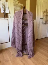 Ruana tejida a mano en color lila, confeccionada en lana merino. Presenta un diseño de punto con calados diagonales y terminación de flecos largos en el ruedo.