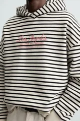 Sudadera con capucha a rayas horizontales blancas y negras, corte boxy fit, mangas largas y texto bordado en color rosa en el pecho.