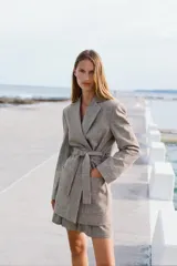 Blazer marrón claro con estampado a cuadros, cuello solapa, manga larga con hombreras, bolsillos delanteros y cierre cruzado con cinturón del mismo tejido.