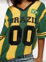 Remerón tejido a crochet con rayas horizontales verdes y amarillas. Tiene cuello en V con ribete negro, mangas cortas con ribete negro y un parche con la bandera de Brasil en el pecho. En el frente tiene bordada la palabra "Brazil" y el número "00" en color negro.