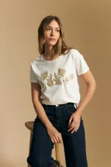 Remera blanca de algodón con cuello redondo y manga corta, con un bordado horizontal de flores estilizadas en tono beige o dorado claro en el centro del pecho.