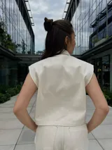 Chaleco color crema con cuello camisero, sin mangas, con cierre frontal de botones a presión y bolsillos de parche en el pecho.