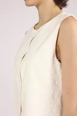 Chaleco negro sin mangas, con cuello redondo y cierre frontal de botones. Presenta detalles de encaje o apliques en los laterales y un corte ligeramente holgado.