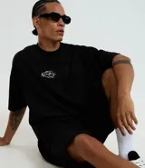 Remera de manga corta, cuello redondo y corte boxy, confeccionada en gamuza sintética (suede) de color negro. Presenta una pequeña estampa localizada en el pecho con un diseño abstracto en tonos dorados/bronce.