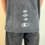 Remera negra con efecto lavado y estampado rectangular con imagen de surfistas en la playa y texto "Visions" en color lila. Corte oversize.