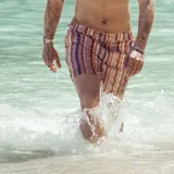 Short de baño con estampado de rayas horizontales y motivos geométricos en tonos naranja, marrón y blanco. Cuenta con pretina elástica y cordón ajustable.