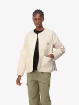 Campera Carhartt Skyler Liner para mujer, color crema, confeccionada en poliéster ripstop reciclado con forro de poliéster acolchado. Presenta cuello redondo, cierre frontal con botones a presión y tres bolsillos delanteros, uno de ellos con etiqueta cuadrada tejida con el logo de la marca.