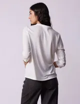 Blusa blanca de poliéster y elastano, con cuello a la base, escote en V y mangas tres cuartos. Presenta dos bolsillos frontales con cierre metálico dorado.