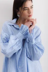 Camisa gris a rayas verticales blancas, de corte amplio, cuello alto con lazo y mangas largas abullonadas con puños elásticos.