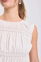 Blusa sin mangas color crudo con escote redondo y frunces horizontales y verticales en el torso, con pespunte contrastante oscuro. Presenta un bajo con volado.