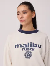 Sweater color ladrillo con cuello redondo en color crema. Estampado frontal con el texto 'Malibu Rusty' y un logo con forma de flor. Corte holgado.