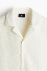 Camisa de manga corta color blanco hueso con textura. Tiene cuello abierto, tapeta lisa y bajo recto.