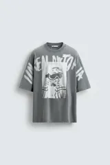 Camiseta gris de corte oversize con cuello redondo y mangas cortas. Presenta un estampado frontal con una fotografía en blanco y negro de una persona con gafas de esquí y texto en aerosol.