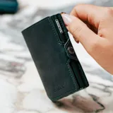 Billetera compacta de cuero genuino con caja de aluminio integrada para tarjetas. Presenta un sistema de deslizamiento mediante clip, bolsillo frontal con cierre de cremallera, compartimento para billetes y documentos, y protección RFID. Diseño en color verde oscuro con costuras a contraste.