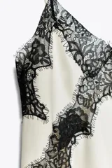 Vestido corto de satén color blanco, con tirantes finos, escote en pico y bajo asimétrico con abertura. Detalle de encaje negro en escote y ruedo.