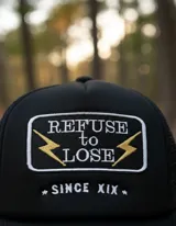 Gorra trucker negra con parche bordado con el texto "Refuse to Lose" y dos rayos dorados.