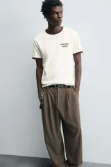 Pantalón negro de corte ancho con cintura elástica, pliegues frontales, bolsillos laterales y traseros de vivo. Tiene detalle de hebilla metálica en la trabilla y cierre frontal con cremallera y botón.
