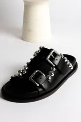 Sandalia tipo mule negra de cuero, con dos tiras ajustables con hebillas metálicas plateadas y apliques de tachas esféricas plateadas. Presenta un borde festoneado en la tira superior y suela de plataforma negra.