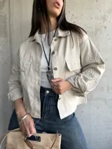 Sobrecamisa color beige con cuello camisero, cierre frontal con botones y bolsillos de parche en el pecho. Presenta detalles de broderie calado en mangas, hombros y bolsillos.