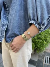 Brazalete rígido dorado con una piedra ovalada de nácar en el centro. Es ajustable y abierto.
