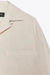 Camisa de manga larga color crudo, confeccionada en hilatura de algodón. Tiene cuello solapa, bolsillo de plastrón en el pecho, bajo redondeado y cierre frontal con botones.