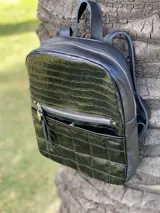 Mochila pequeña negra con textura tipo cuero de cocodrilo en la parte superior y diseño acolchado en la parte inferior. Tiene un bolsillo frontal con cierre, correas ajustables y asa superior.